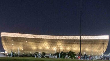 Golden bowl-shaped Lusail Stadium glows at night behind palms and roadway lights.