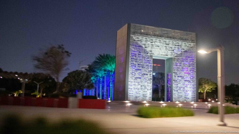 Illuminated rectangular stone portal stands amid landscaped palms and colored night lighting.