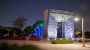 Illuminated rectangular stone portal stands amid landscaped palms and colored night lighting.