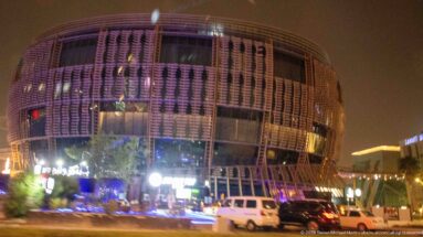 Rounded modern building with a perforated, finned façade glows above shops and parked vehicles at night.