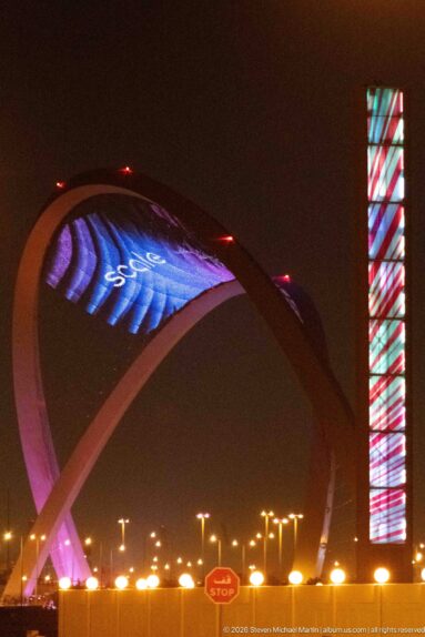 Oblique night view of the pink 5/6 Arch beside a tall illuminated rectangular roadside tower.