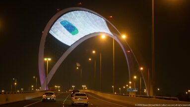 Cars drive toward the illuminated 5/6 Arch spanning a highway beneath a large digital display.