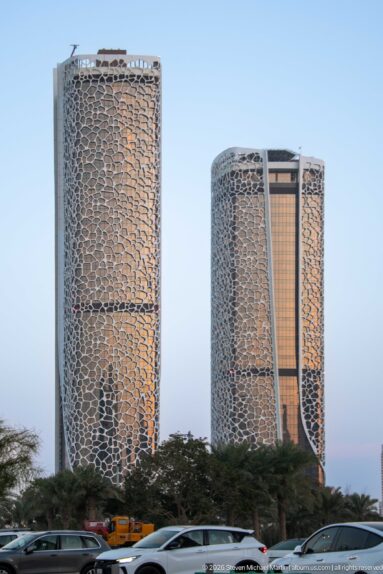 Two rounded high-rises with intricate cellular screen façades rise above trees and traffic at dusk.