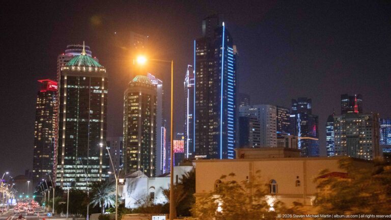 Illuminated Doha high-rises with varied crowns and vertical lights rise behind lower buildings and palms.