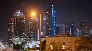 Illuminated Doha high-rises with varied crowns and vertical lights rise behind lower buildings and palms.