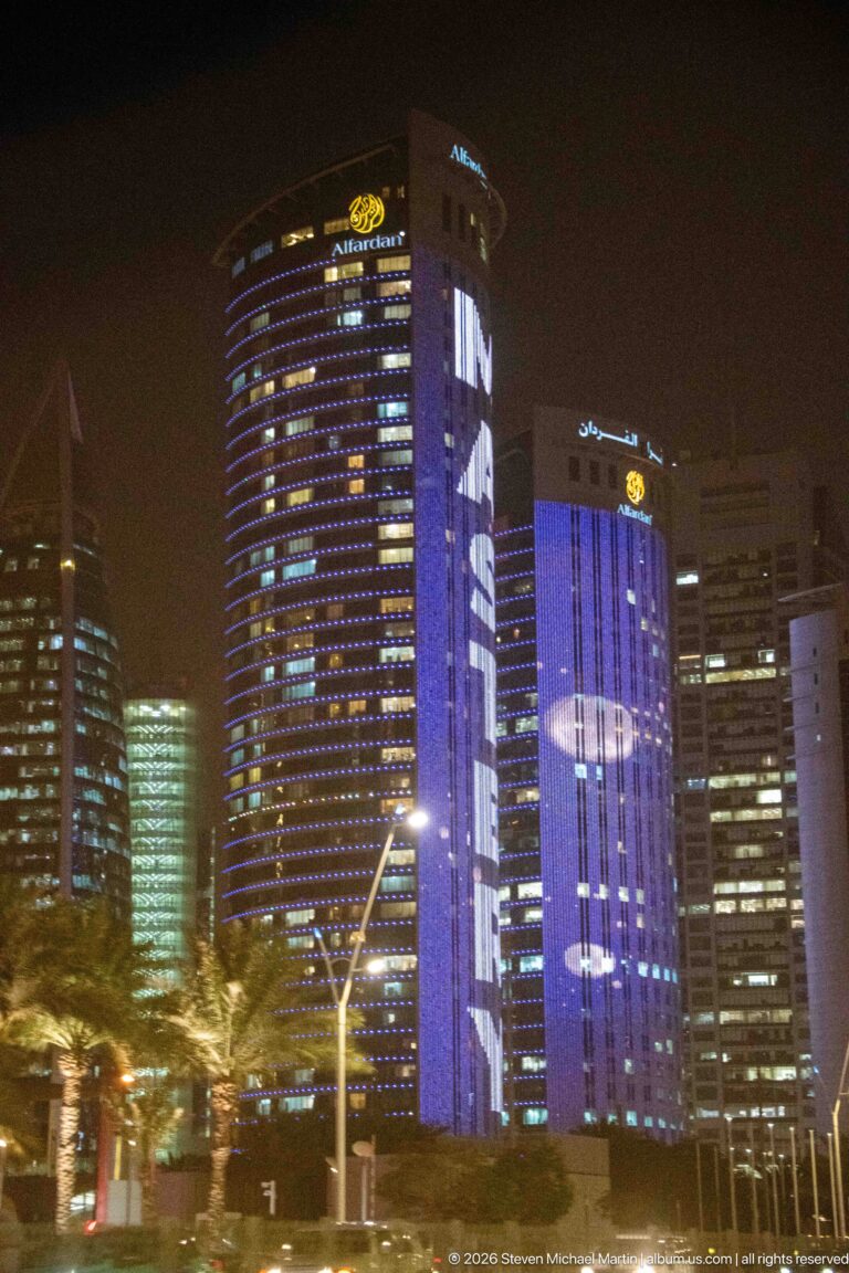 Curved glass towers display large purple and blue illuminated graphics above palms and streetlights at night.