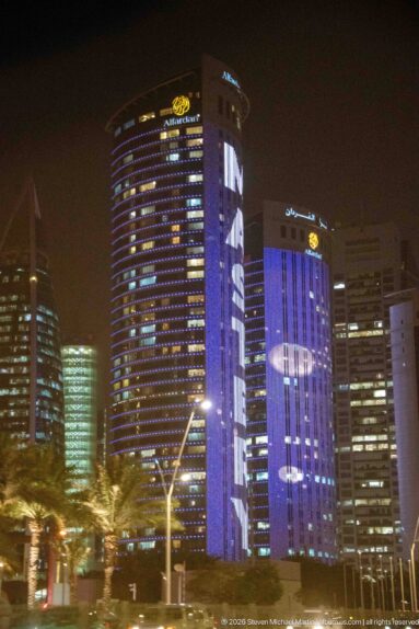 Curved glass towers display large purple and blue illuminated graphics above palms and streetlights at night.