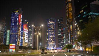 Wide nighttime boulevard leads toward Tornado Tower and brightly illuminated skyscrapers in Doha's West Bay.
