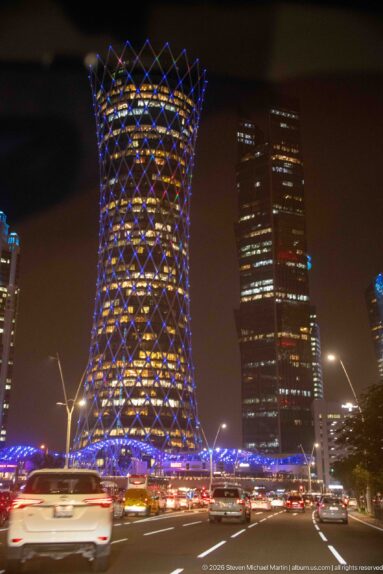 Blue-lit Tornado Tower with diamond lattice rises above nighttime traffic and neighboring skyscrapers in West Bay.