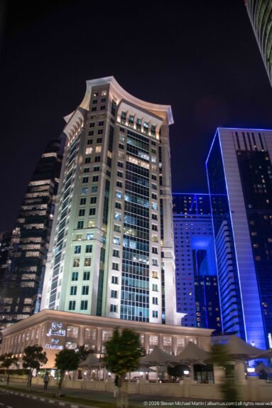 Large pale high-rise with a projecting cornice stands among blue-lit modern towers at night.