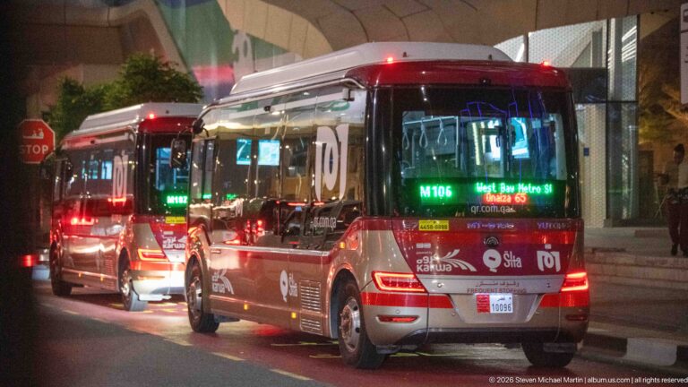 Two red-and-silver Karwa buses wait in nighttime traffic beside a modern building.