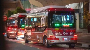 Two red-and-silver Karwa buses wait in nighttime traffic beside a modern building.
