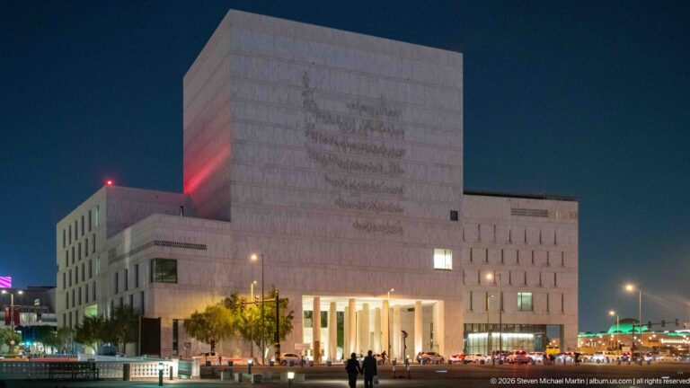 Large pale stone institutional building with a tall recessed colonnaded entrance illuminated at night.