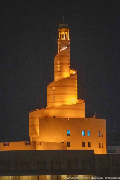 Close night view of Fanar's amber-lit spiral minaret and stepped base against a black sky.