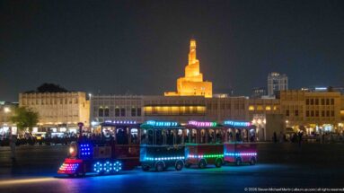 Colorful illuminated tourist road train passes in front of Fanar's golden spiral minaret at night.