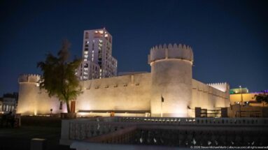 Warmly lit Al Koot Fort with a round corner tower and crenellated walls, backed by a modern high-rise.