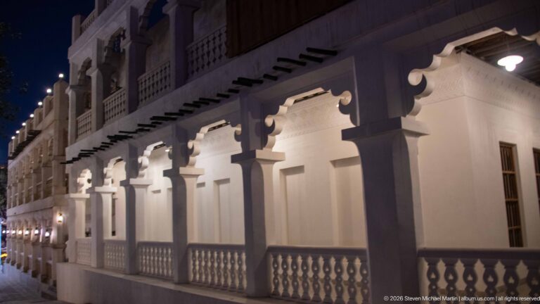 White colonnade with decorative brackets, projecting beams, and a low balustrade illuminated at night.