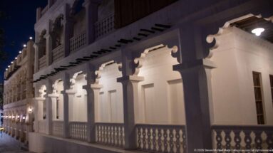 White colonnade with decorative brackets, projecting beams, and a low balustrade illuminated at night.