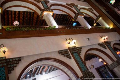 Decorated arches, turquoise geometric panels, timber ceiling beams, and colorful hanging lamps fill a traditional-style interior.