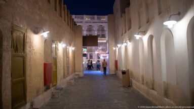 Narrow paved alley between weathered walls and white arches, lit by wall lamps at night.