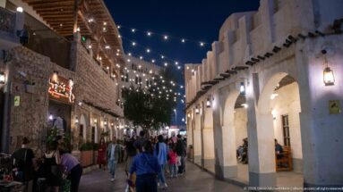 Pedestrians walk beneath string lights in a narrow lane between stone shops and a whitewashed arcade.
