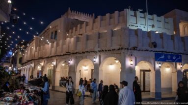 White arcaded market building with stepped crenellations stands beneath strings of lights and evening crowds.