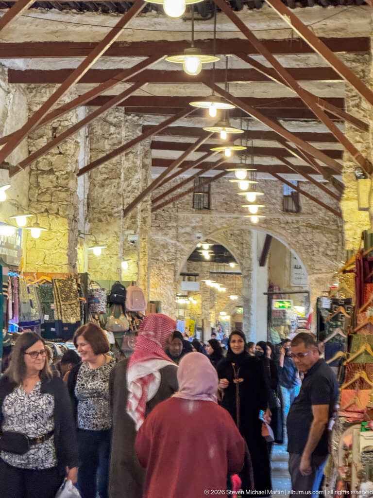 Crowds fill a covered stone market aisle lined with textiles and merchandise under a metal roof.