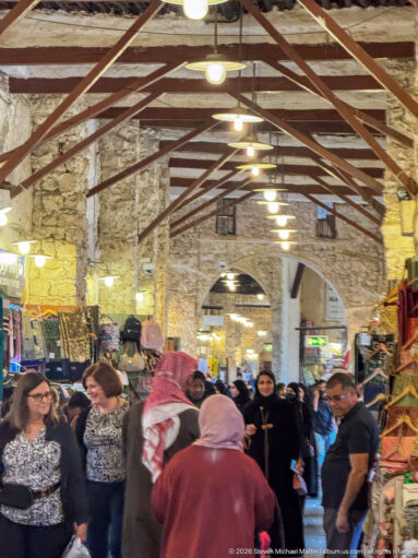 Crowds fill a covered stone market aisle lined with textiles and merchandise under a metal roof.
