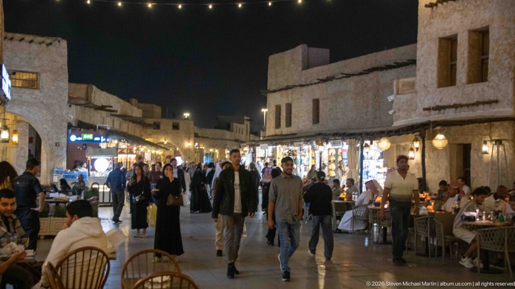 People walk between restaurant tables and illuminated shops along a narrow Souq Waqif lane at night.