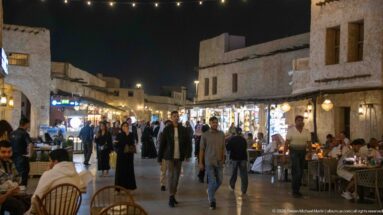 People walk between restaurant tables and illuminated shops along a narrow Souq Waqif lane at night.