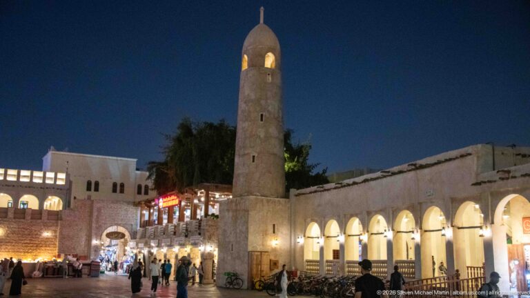 Stone minaret with a domed top rises beside a brightly lit arcade and busy pedestrian lane at night.