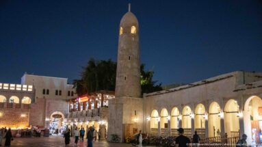 Stone minaret with a domed top rises beside a brightly lit arcade and busy pedestrian lane at night.
