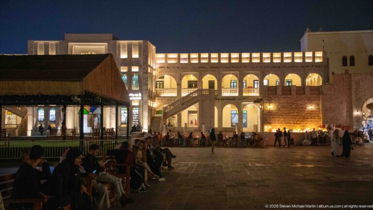 People sit around a paved square bordered by illuminated arcades, balconies, and café seating at night.