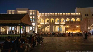 People sit around a paved square bordered by illuminated arcades, balconies, and café seating at night.