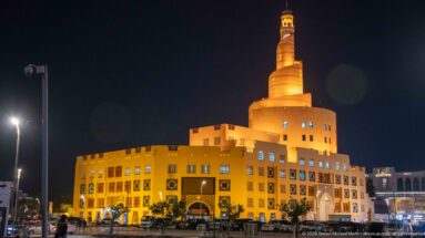 Amber-lit spiral minaret rises above the broad base of the Fanar Islamic Cultural Center at night.