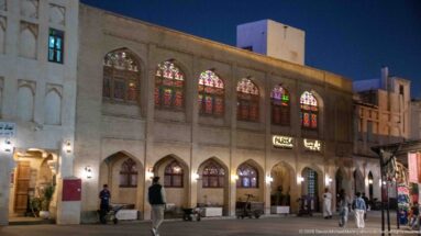Pedestrians pass a two-story market building with pointed arches and glowing colored-glass windows at night.
