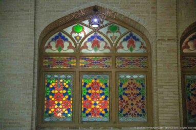 Pointed-arch window filled with brightly colored geometric and floral stained glass in a pale masonry wall.
