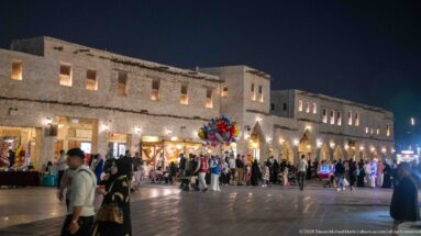 Crowded paved plaza outside illuminated Souq Waqif buildings at night, with glowing shops and a cluster of balloons.