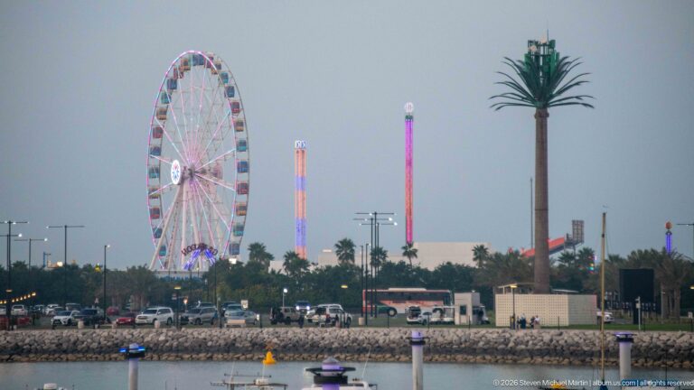 Illuminated Ferris wheel and tall amusement rides stand beyond a calm waterfront at dusk.