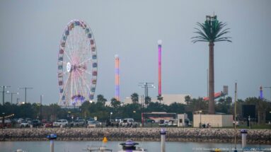 Illuminated Ferris wheel and tall amusement rides stand beyond a calm waterfront at dusk.