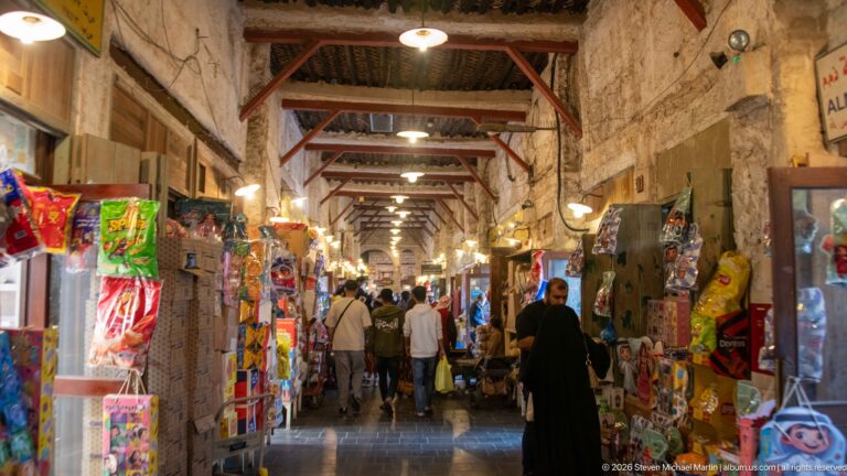 Covered Souq Waqif aisle lined with colorful packaged goods and small shops, with shoppers walking through the narrow passage.