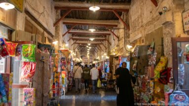 Covered Souq Waqif aisle lined with colorful packaged goods and small shops, with shoppers walking through the narrow passage.