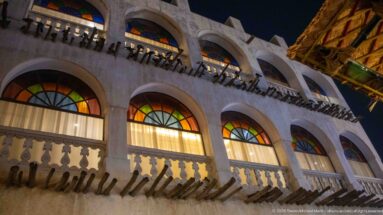 Upward view of a Souq Waqif façade with arched colored-glass windows, pale balustrade, and projecting timber beams at night.