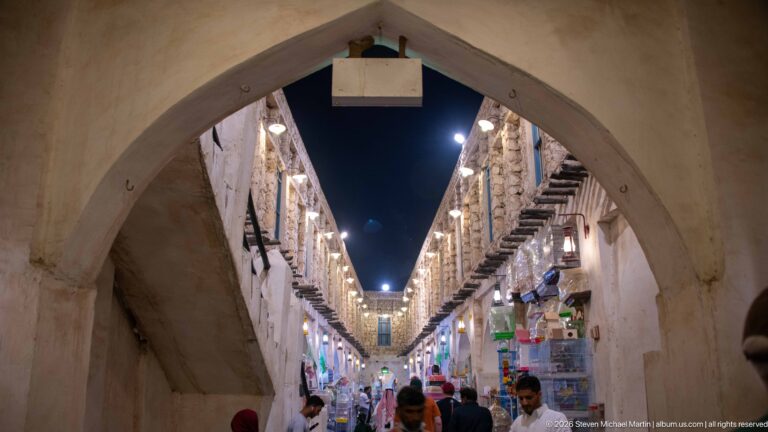 Pointed arch opening onto a crowded Souq Waqif lane lined with shops, projecting wooden beams, and rows of lights.