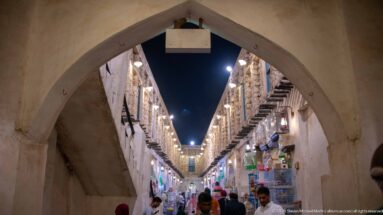 Pointed arch opening onto a crowded Souq Waqif lane lined with shops, projecting wooden beams, and rows of lights.