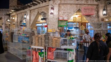 Souq Waqif shop beneath rough plaster arches, with stacked packaged goods, hanging lights, shoppers, and a vendor.