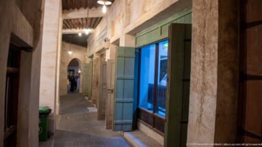 Narrow Souq Waqif passage with rough plaster walls, turquoise shutters, a blue-lit window, and small lamps receding into the distance.