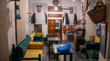 Green wooden benches with yellow cushions, low tables, portraits, baskets, and a model dhow in a traditional interior.