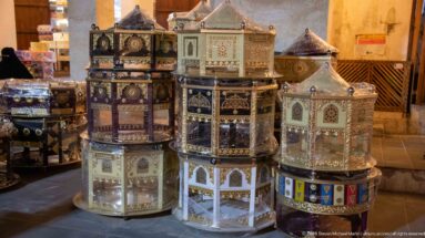 Stacked decorative metal lanterns with colored glass, pierced panels, geometric designs, and small domed tops in a market display.