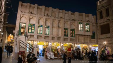 Nighttime Souq Waqif lane with traditional shopfronts, green-trimmed windows, open stores, and pedestrians gathered outside.
